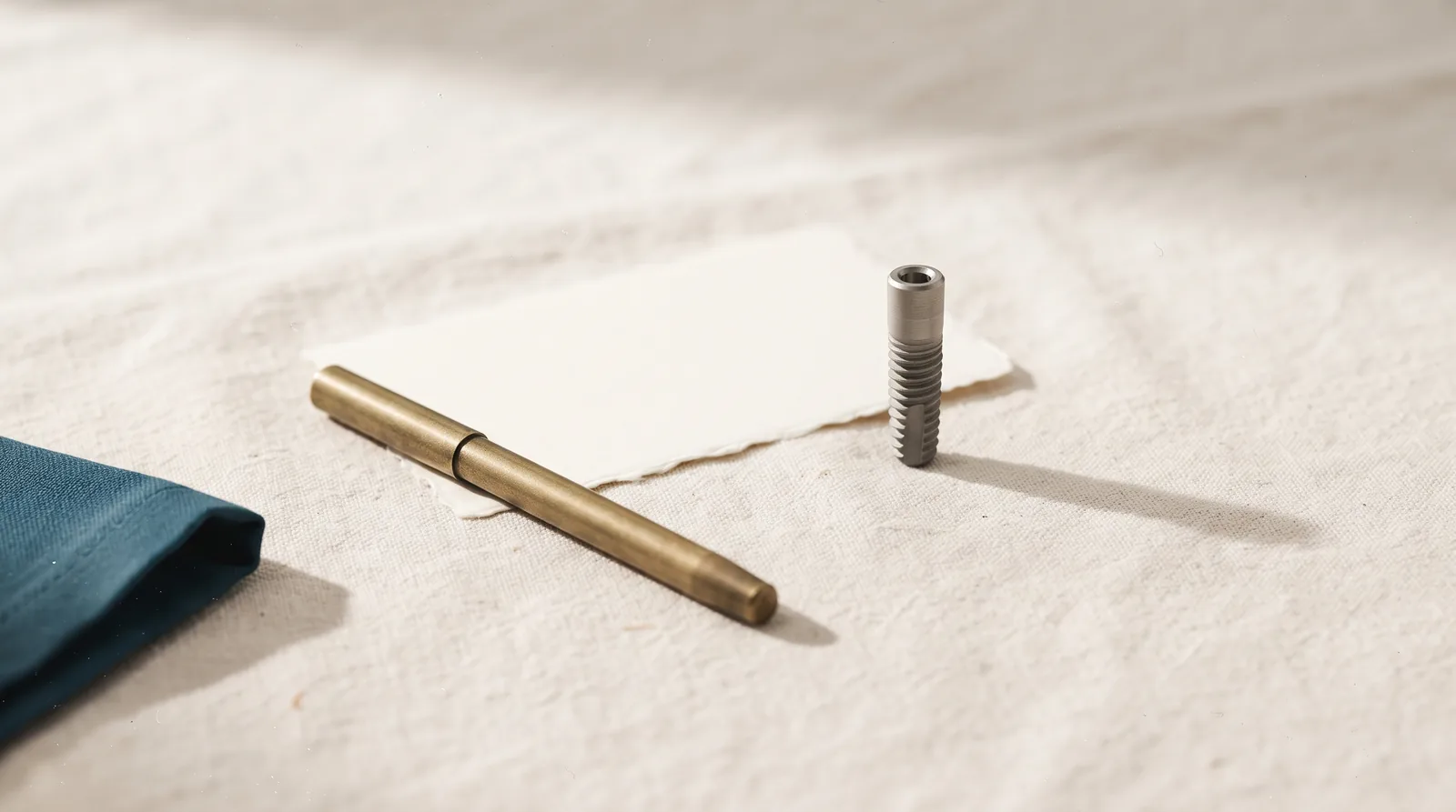 A titanium dental implant post and a blank ivory notecard with a brass pen on linen
