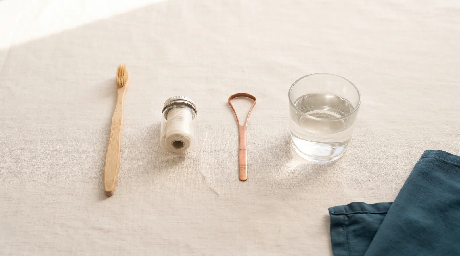 A toothbrush, floss, tongue scraper and rinse glass laid in a row on linen