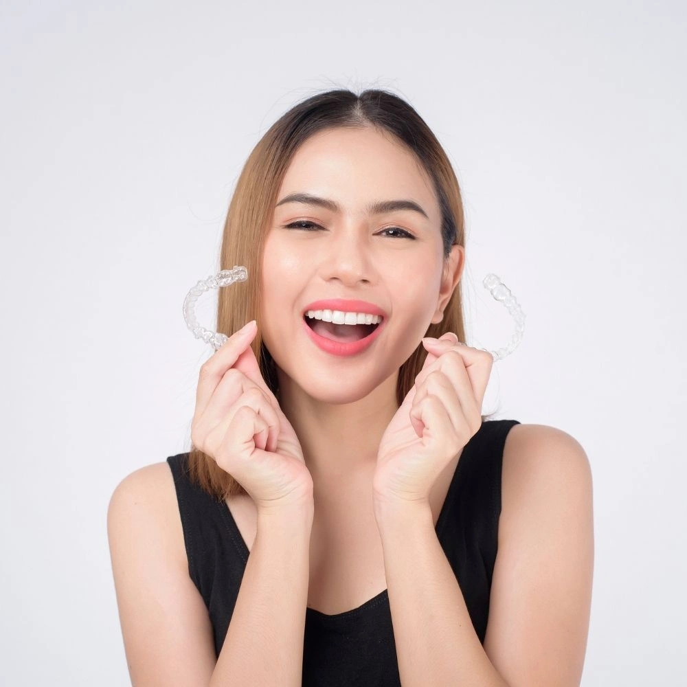 A young woman happily holding clear aligners near her teeth, showcasing her bright smile. Promotional image for clear aligner treatment at Dental Art Clinic.