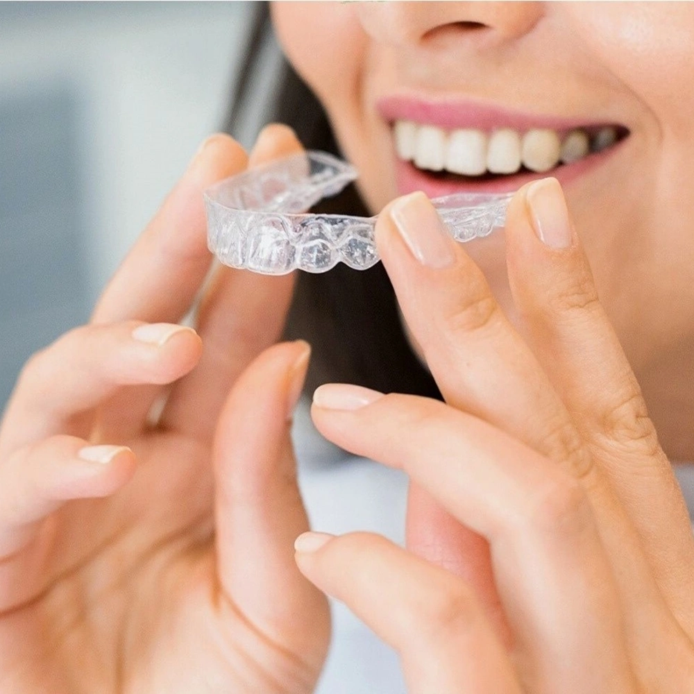 A woman holding and placing a clear dental aligner on her upper teeth, showing a close-up view of her smile and the transparent orthodontic appliance.